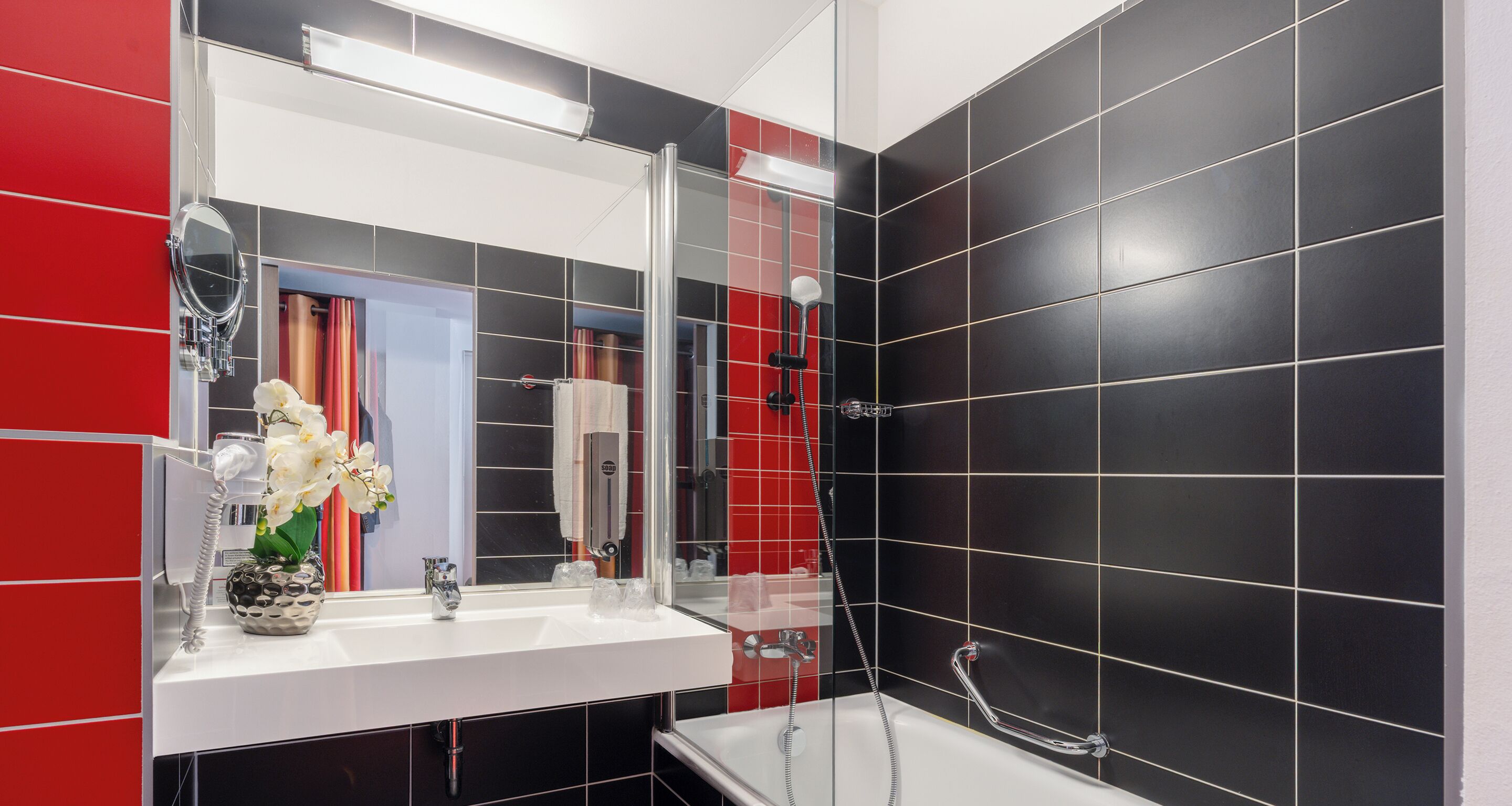 Une salle de bain moderne avec un lavabo blanc, une douche en verre et des murs recouverts de carreaux noirs et rouges.