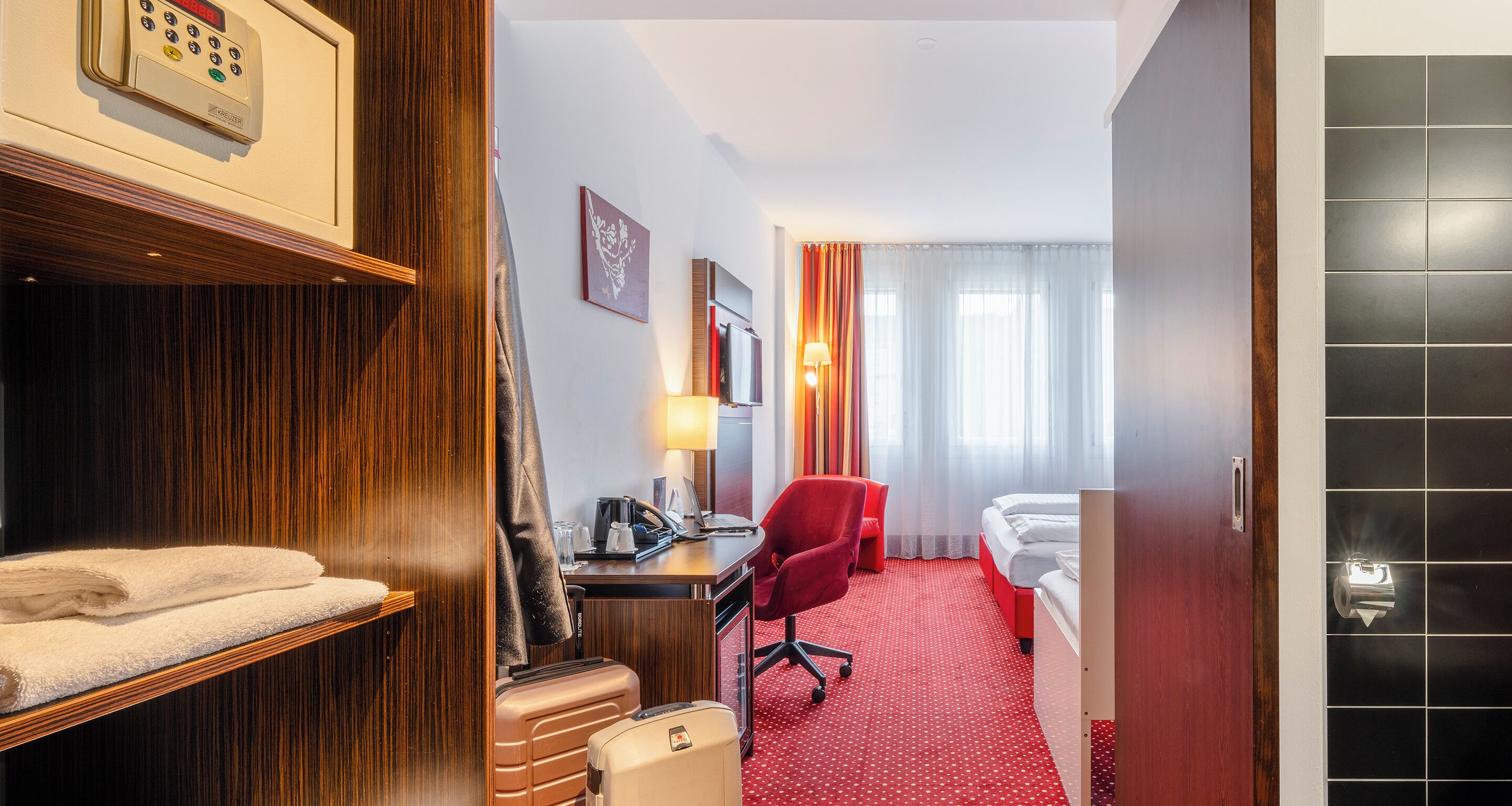 A hotel room with a wooden wardrobe, a desk with a lamp, a red chair, a bed, and a bathroom with a towel rack.