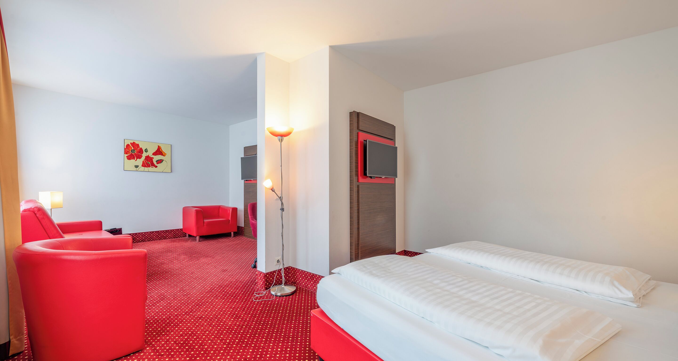 Habitación de hotel con alfombra roja, una cama con ropa de cama blanca, un sillón rojo, un sofá rojo, una lámpara de pie y un cuadro en la pared.