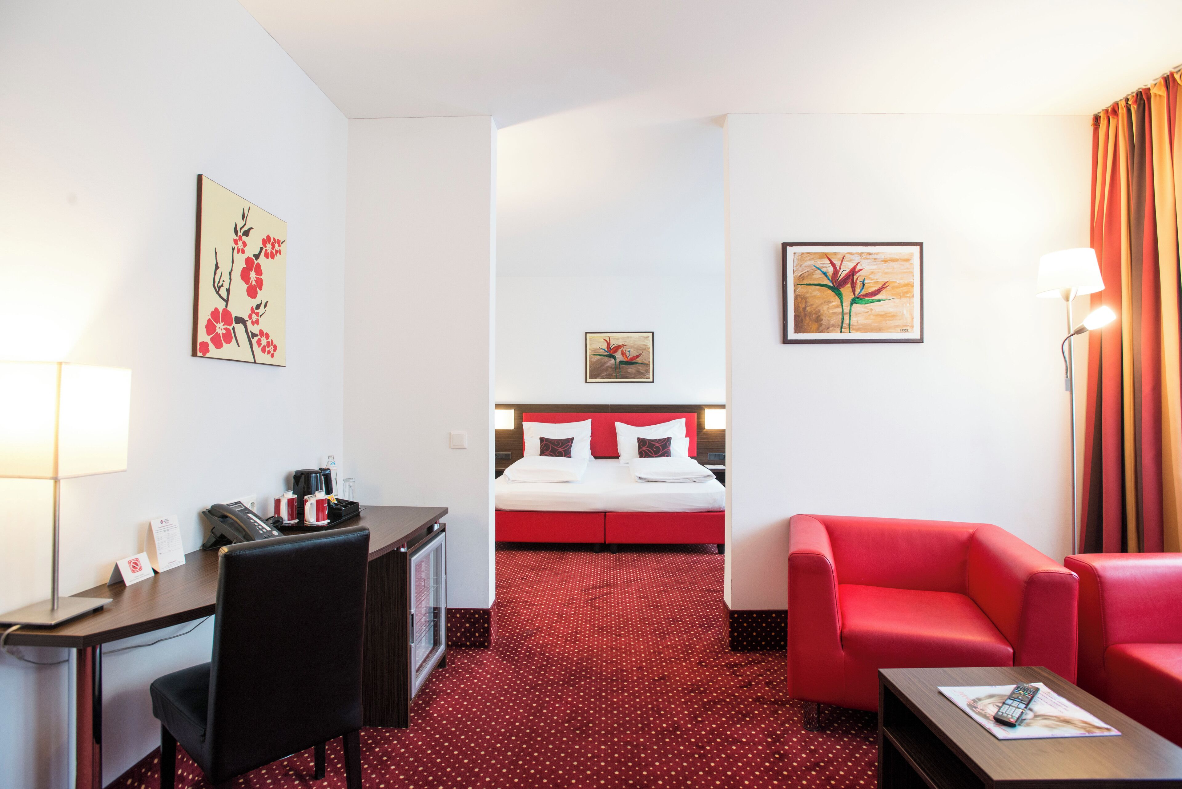 Quarto de hotel com cama, escrivaninha, cadeira, sofá vermelho e obras de arte nas paredes.