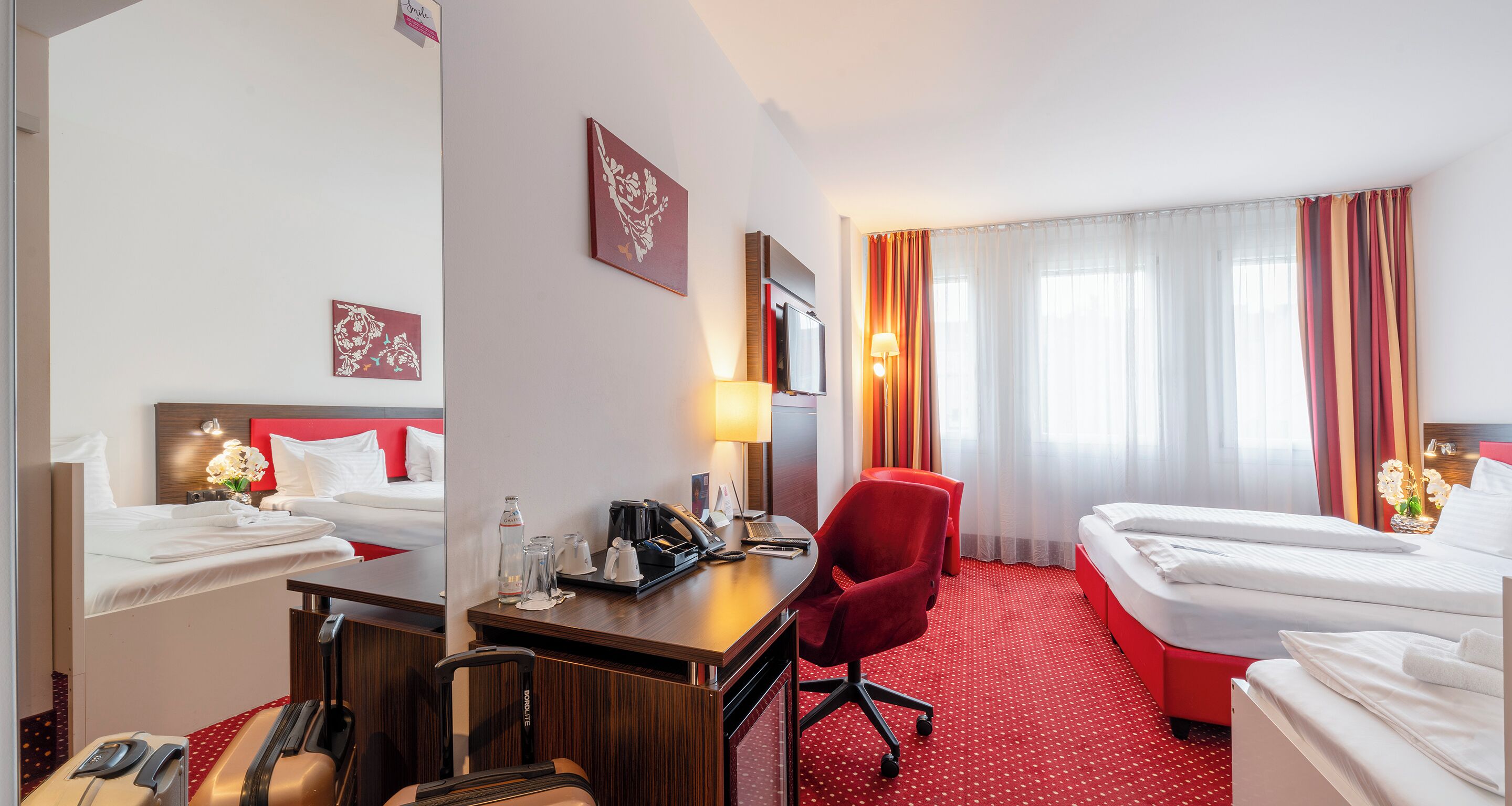 A hotel room with two beds, a desk with a lamp, a chair, and a suitcase.
