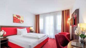 Una habitación de hotel con una cama roja, almohadas blancas, una silla roja, un escritorio con una computadora portátil y un jarrón con flores.
