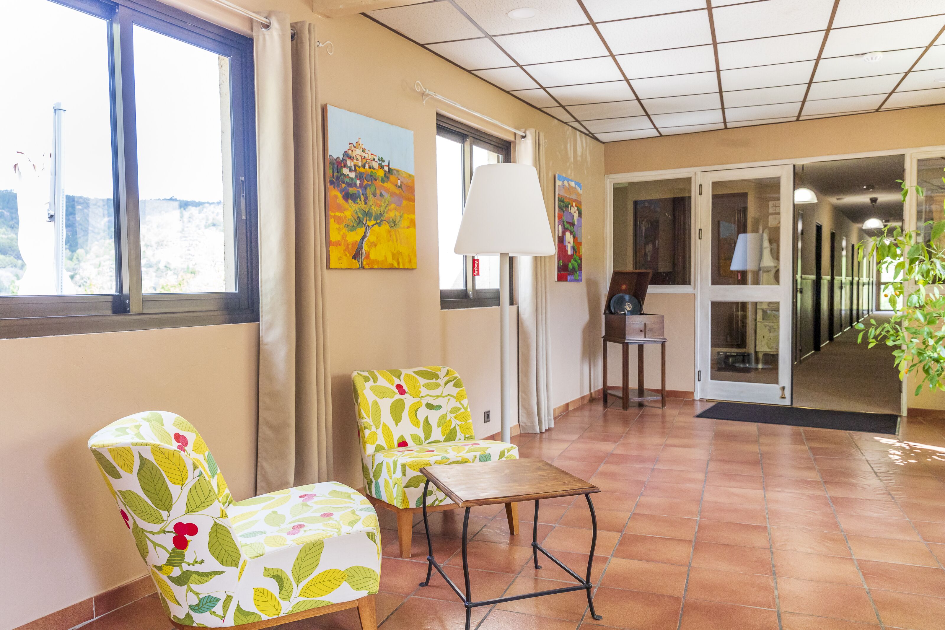 Um saguão de hotel com piso de azulejos, dois poltronas estampadas, uma mesa pequena e um quadro na parede.
