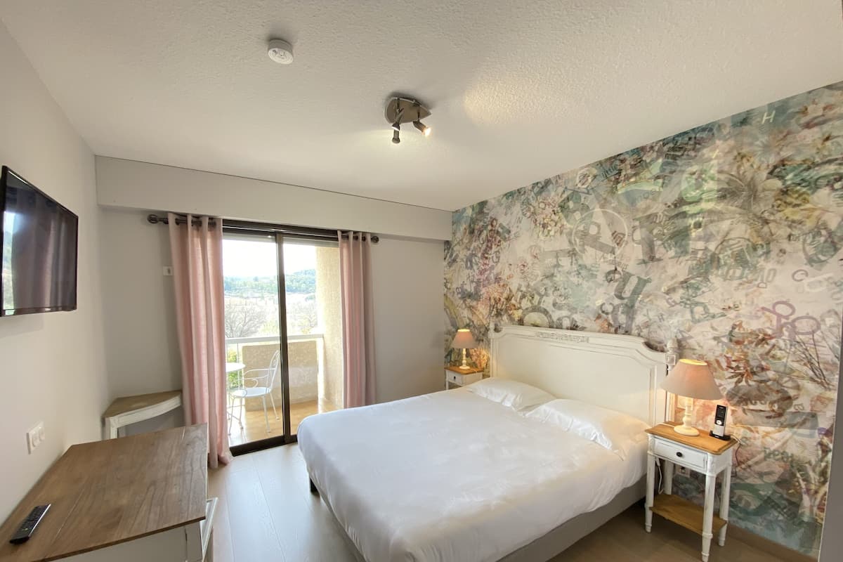 A hotel room with a large bed, a desk, a chair, a small table with a lamp, and a view of the outdoors through a sliding door.