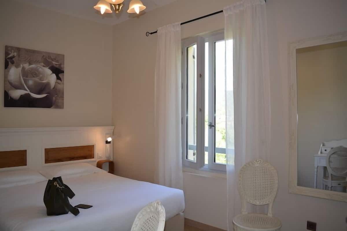 A hotel room with a bed, a chair, a window with curtains, a wall-mounted lamp, and a painting.