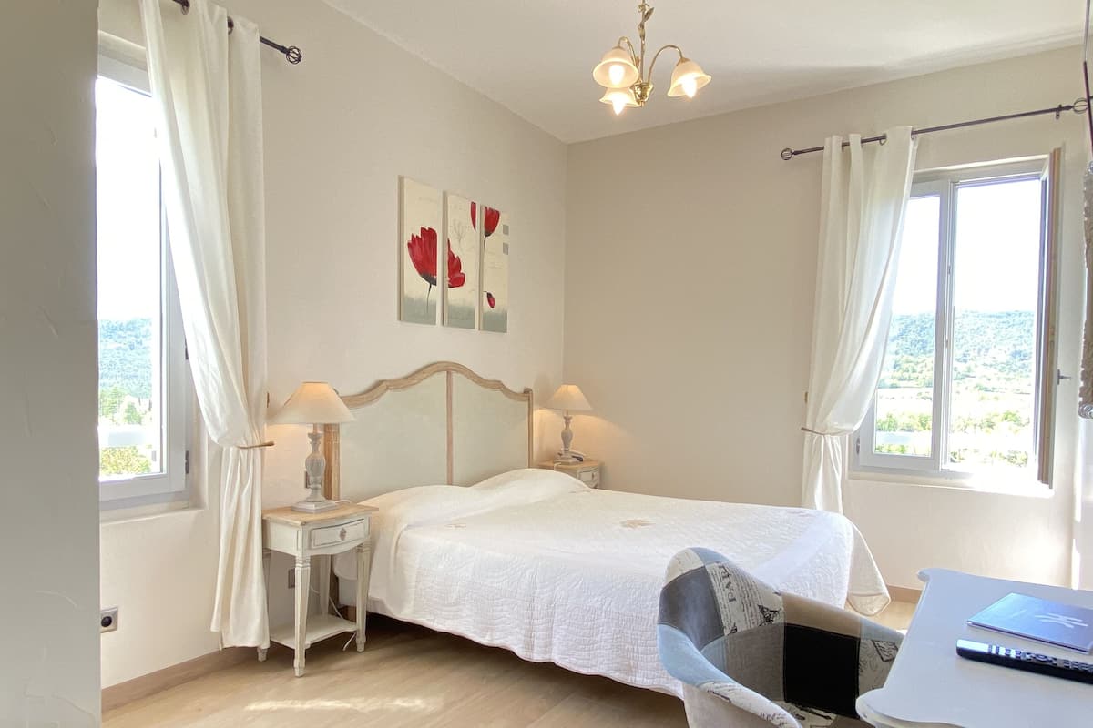 A bedroom with a bed, two bedside tables with lamps, a chair, a desk with a laptop, and a view of the outdoors through the window.