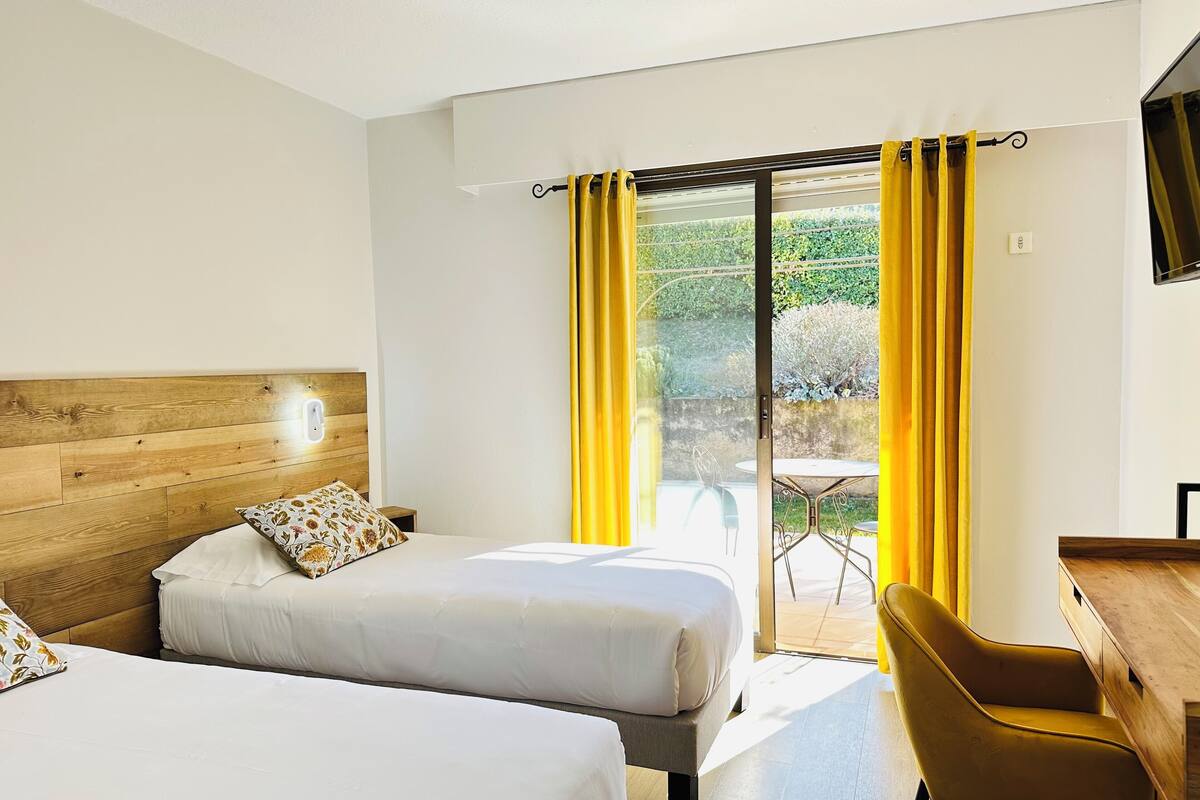 A hotel room with two beds, a wooden headboard, a desk, a chair, and a sliding glass door leading to an outdoor area with a table and chairs.