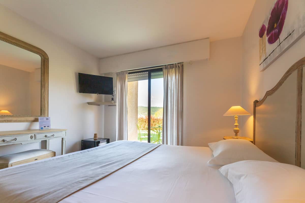 A hotel room with a bed, a desk with drawers, a television mounted on the wall, and a window with curtains.