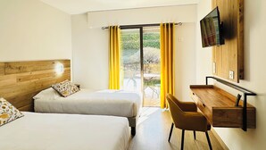 A hotel room with two beds, a desk with a chair, a TV mounted on the wall, and a sliding glass door leading to an outdoor area. - Hôtel Le Colombier (Moustiers-Sainte-Marie)