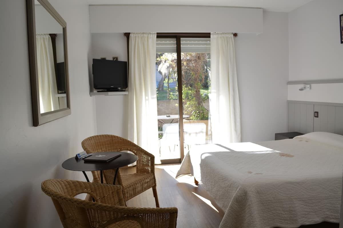 A hotel room with a bed, a television, a wicker chair, a small table, and a balcony with curtains.
