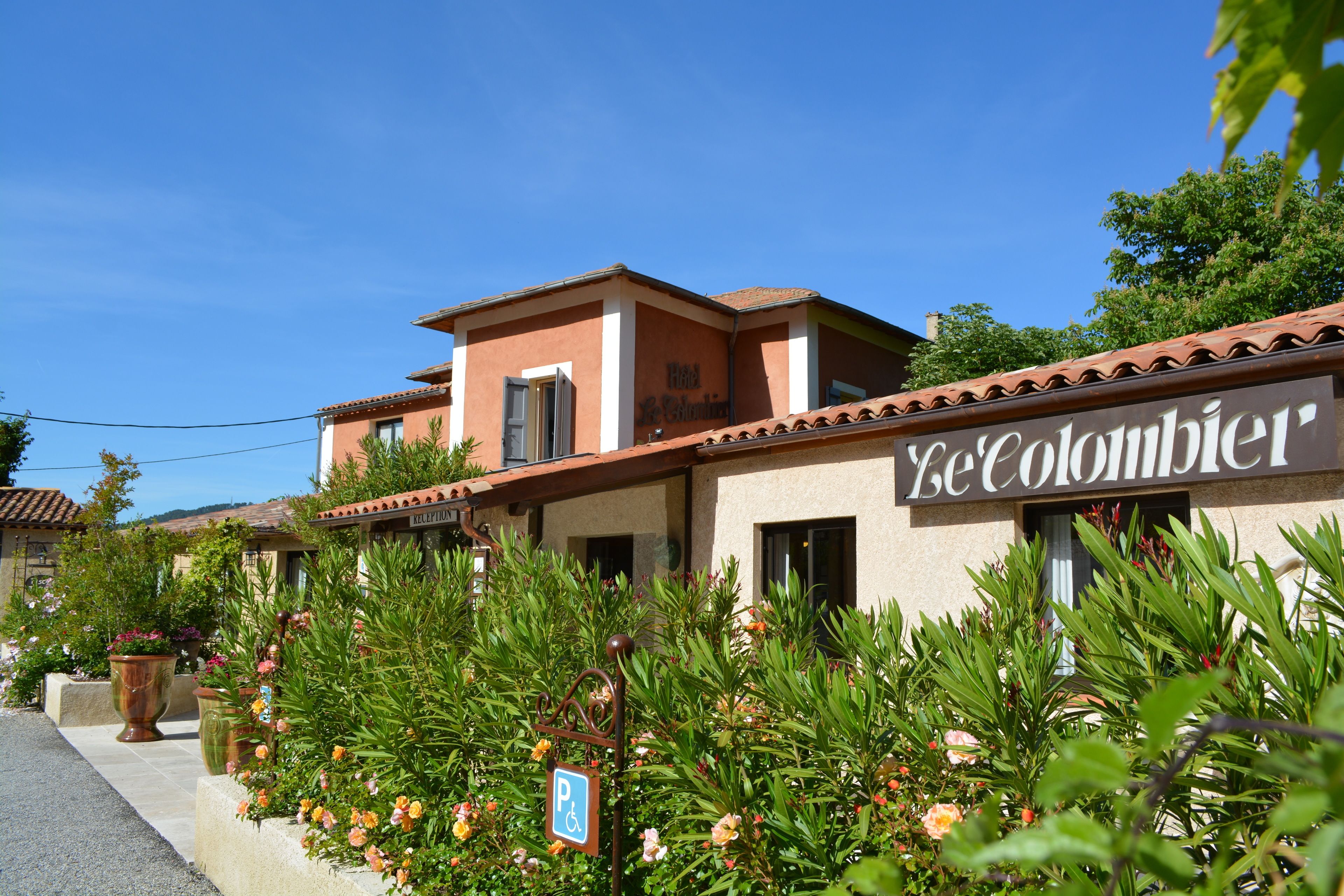 Hotel anomenat "Hotel Le Colombier" amb un rètol, un senyal de pàrquing i vegetació abundant al davant.
