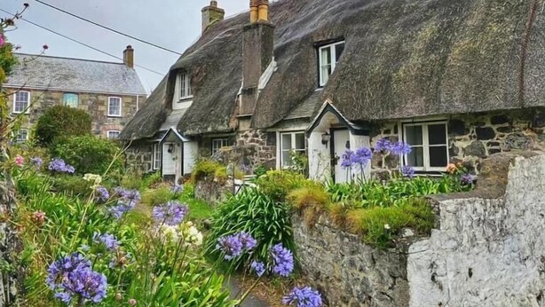 A row of traditional stone houses with thatched roofs, surrounded by lush gardens and flowering plants.