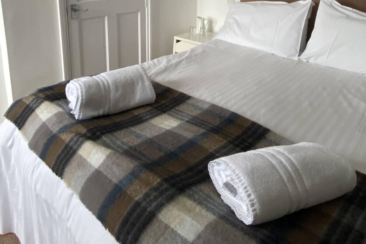 A neatly made bed with a plaid blanket and two rolled towels, a white door, and a nightstand with a lamp.