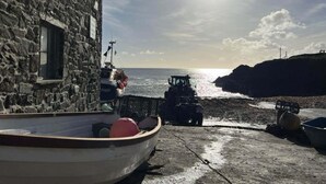 A stone building with a boat docked in front, a tractor on the beach, and a cross on a hill in the background.