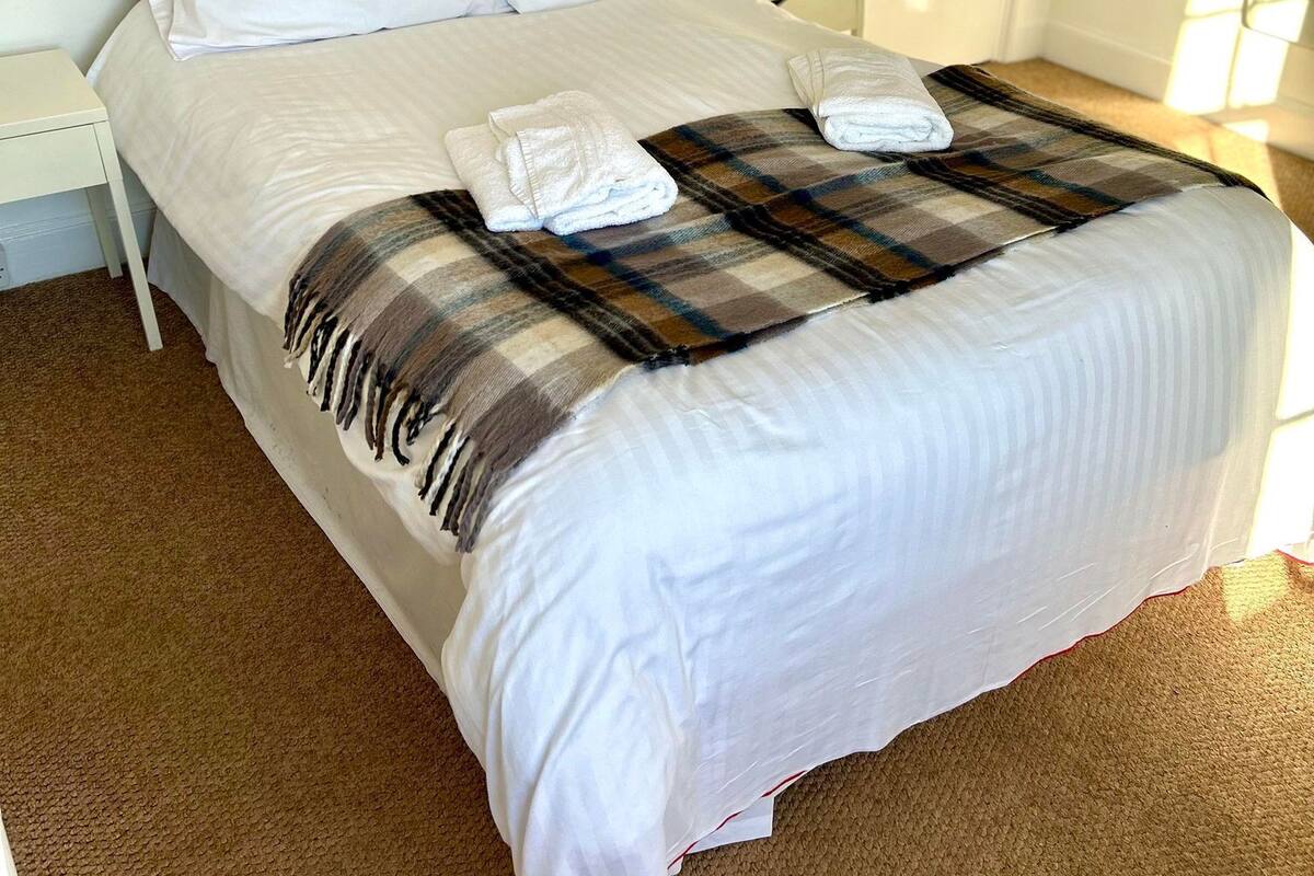 A neatly made bed with white linens and a plaid blanket, a bedside table with a lamp, and a door with a coat rack in a well-lit room.