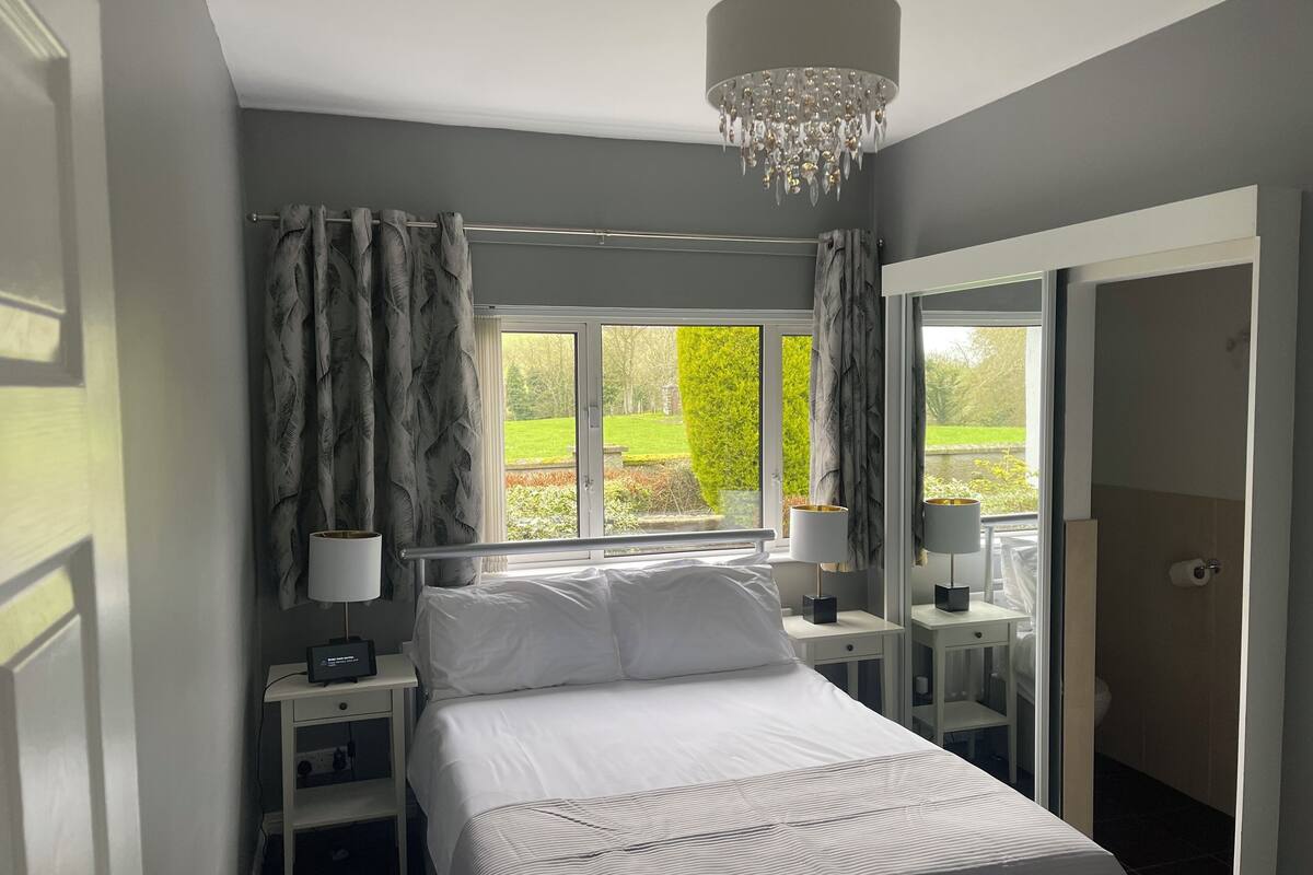 Une chambre à coucher avec un lit, deux tables de chevet avec des lampes, un lustre, une fenêtre avec des rideaux et une vue sur la verdure à l’extérieur.