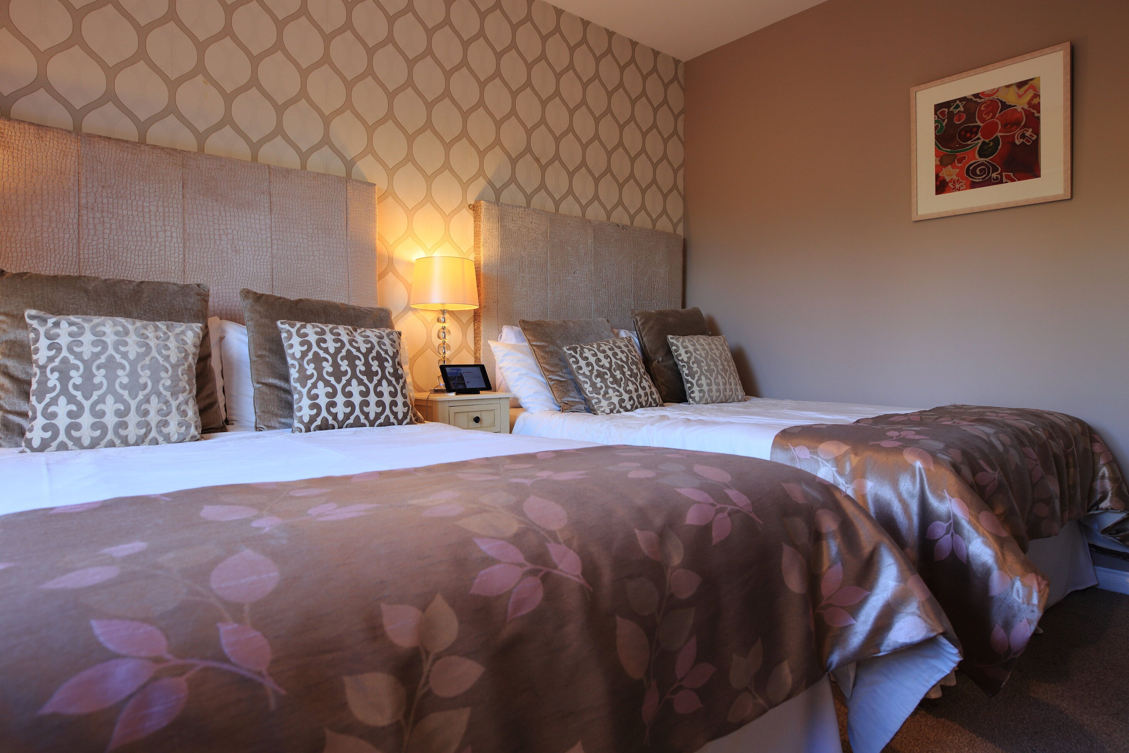 A hotel room with two beds, patterned pillows, a nightstand with a lamp, and a framed picture on the wall.