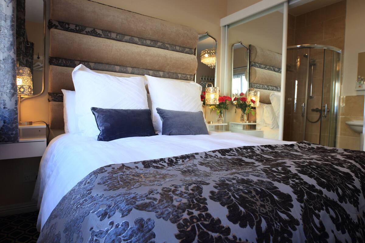 Une chambre d’hôtel avec un grand lit, une table de chevet, un miroir, un espace douche et un vase avec des fleurs.