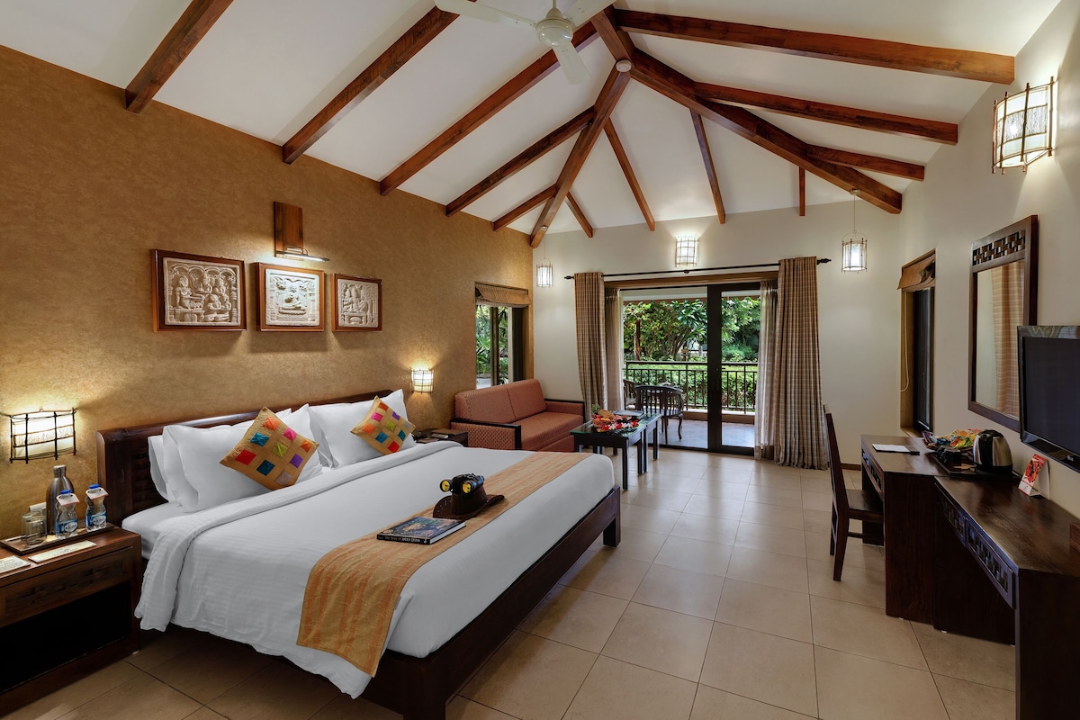 A hotel room with a bed, a desk, a television, and a balcony with a view of greenery.