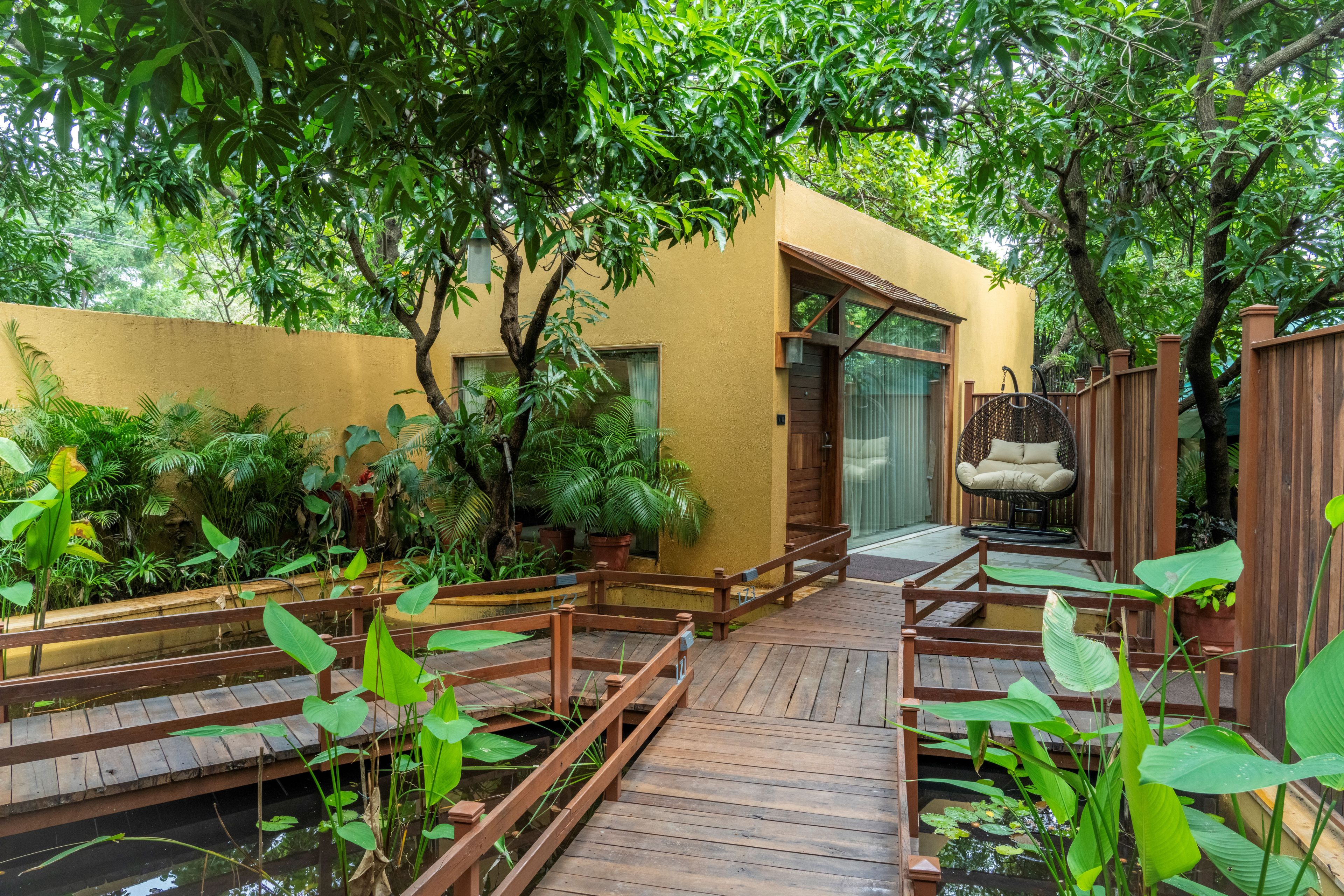A wooden deck leading to a yellow building with a glass door, surrounded by lush greenery and a small pond.