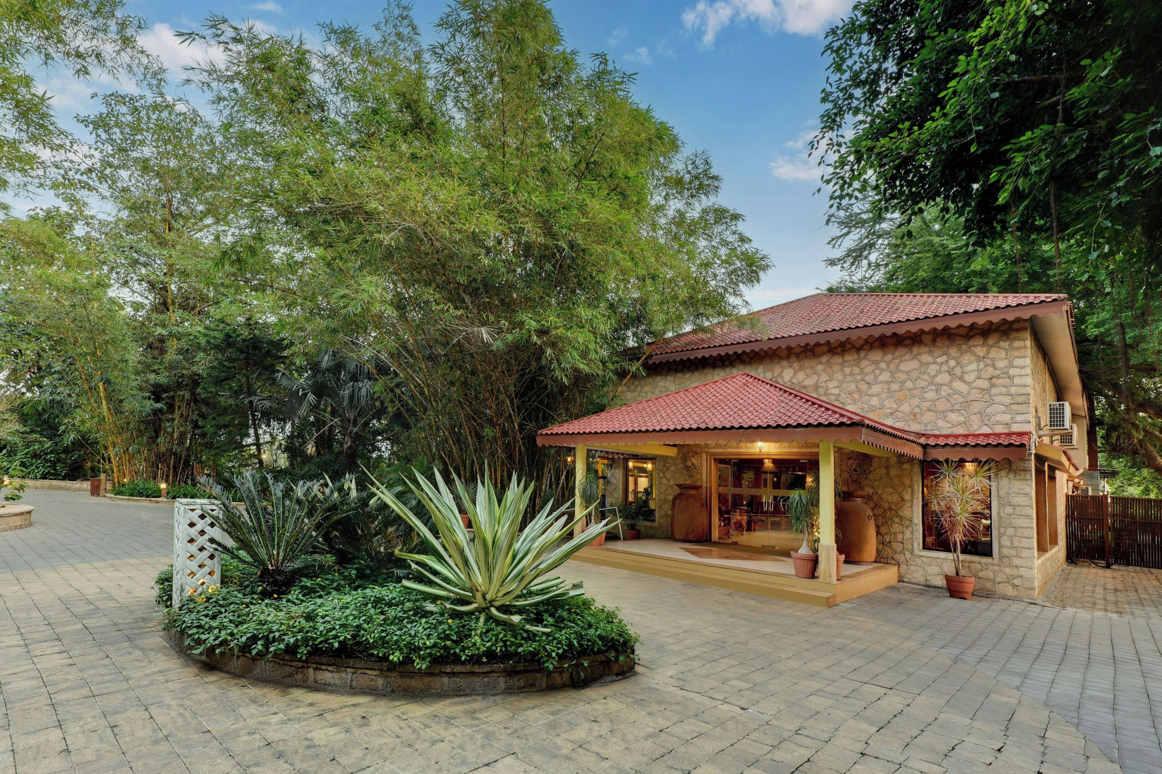 A stone building with a red-tiled roof, surrounded by lush greenery and a paved entrance area.
