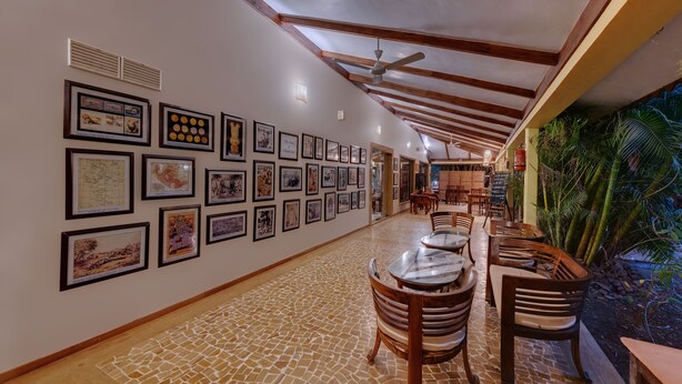 A corridor with framed artworks on the walls, wooden tables and chairs, and a ceiling fan. - The Fern Gir Forest Resort Sasan Gir, Series by Marriott (Mendarda)