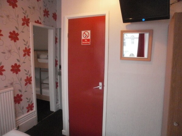 Habitación de hotel con puerta roja, cartel de prohibido fumar, un espejo pequeño y un televisor montado en la pared.