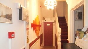 Un pasillo con una escalera que sube a un piso superior, una puerta roja, una lámpara fijada a la pared y un cuadro enmarcado.