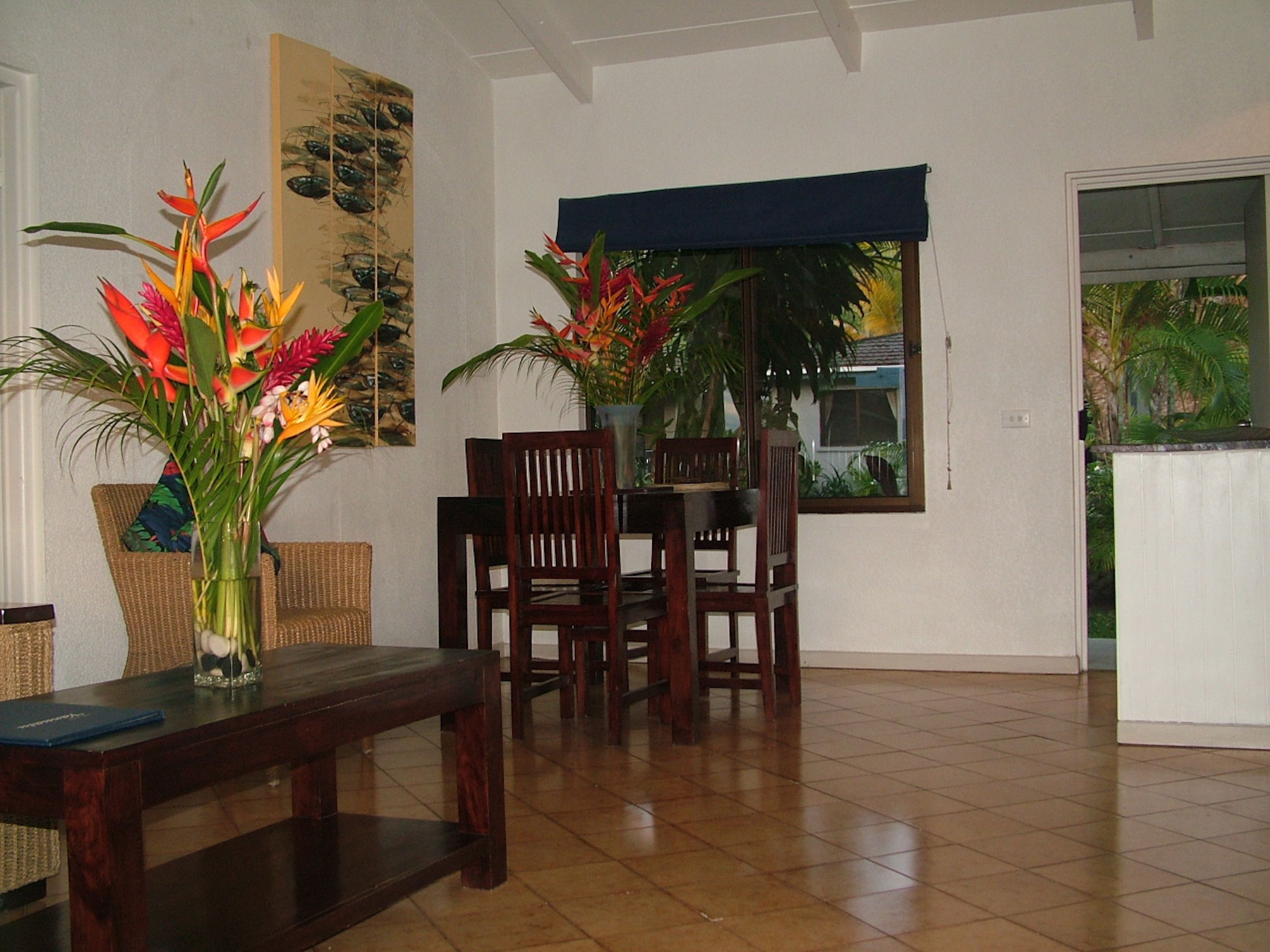 A dining area with a wooden table and chairs, a vase with tropical flowers, and a window with a view of greenery.