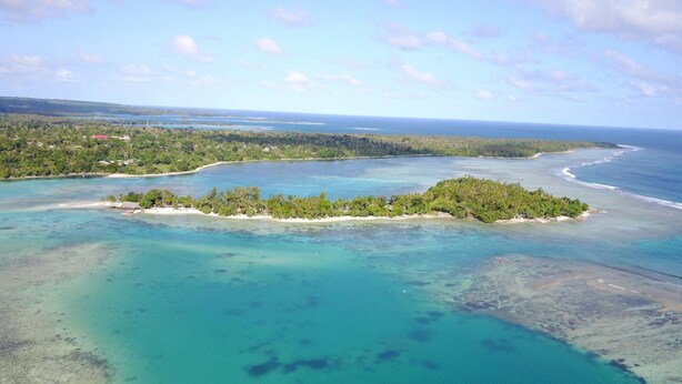 Vista aérea de una isla tropical con aguas turquesas cristalinas, rodeada de exuberante vegetación y una costa distante.