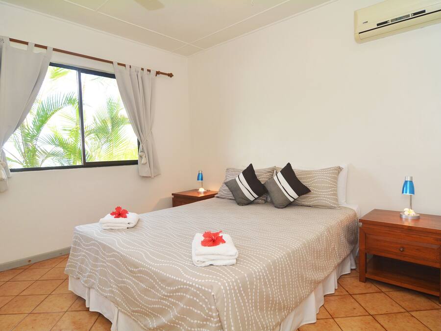 A neatly made bed with a white and beige bedspread, two pillows, and two folded towels with a red flower. There are two bedside tables with lamps, a window with curtains, and an air conditioning unit on the wall.