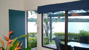 Una habitación con un ventanal grande con vistas a un lago, un comedor con sillas y una mesa, y una maceta con flores anaranjadas y rojas.