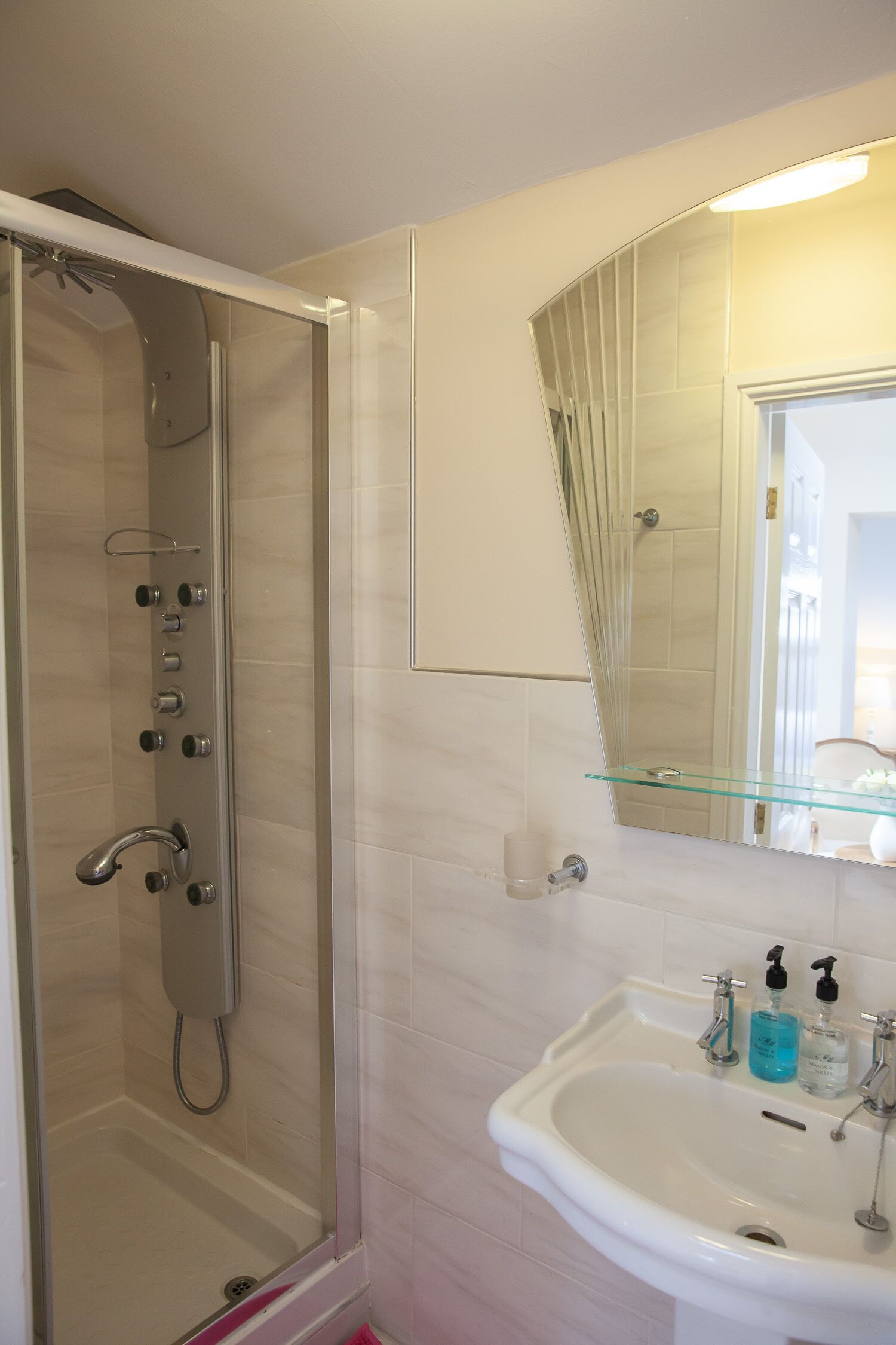 Une salle de bain avec une douche à parois de verre, un lavabo blanc doté d’une étagère en verre et un miroir au-dessus du lavabo.