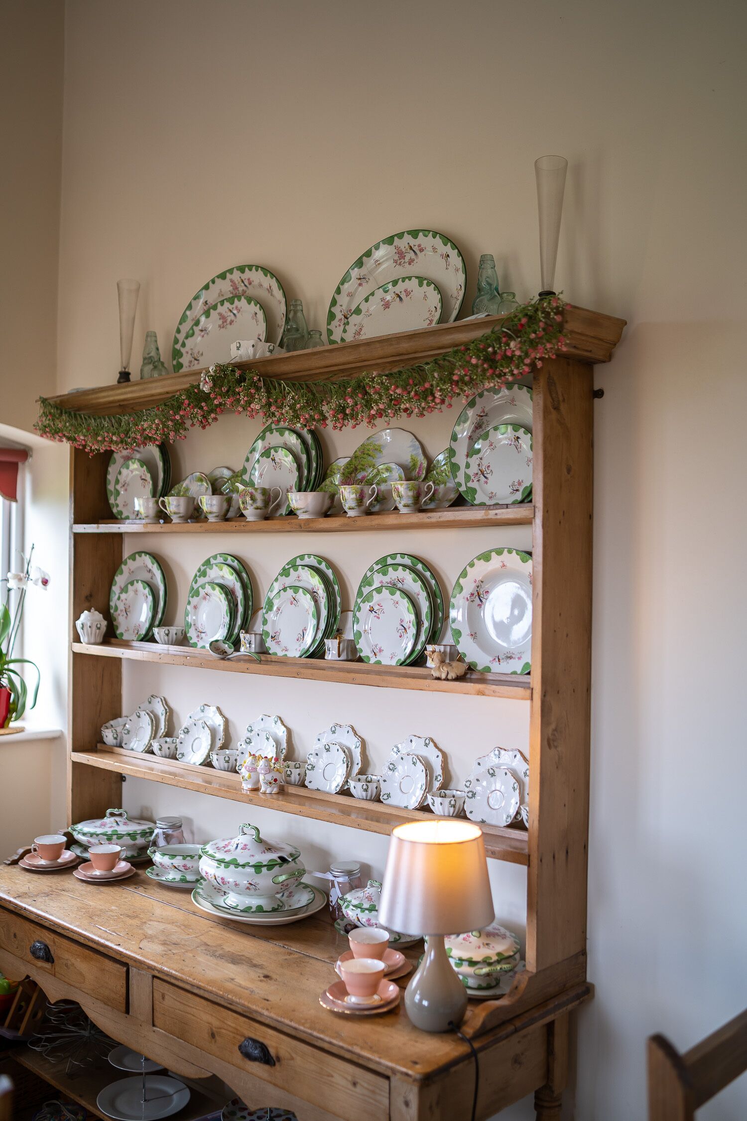 A wooden shelf unit with multiple shelves displaying various ceramic dishes and a table with a lamp.