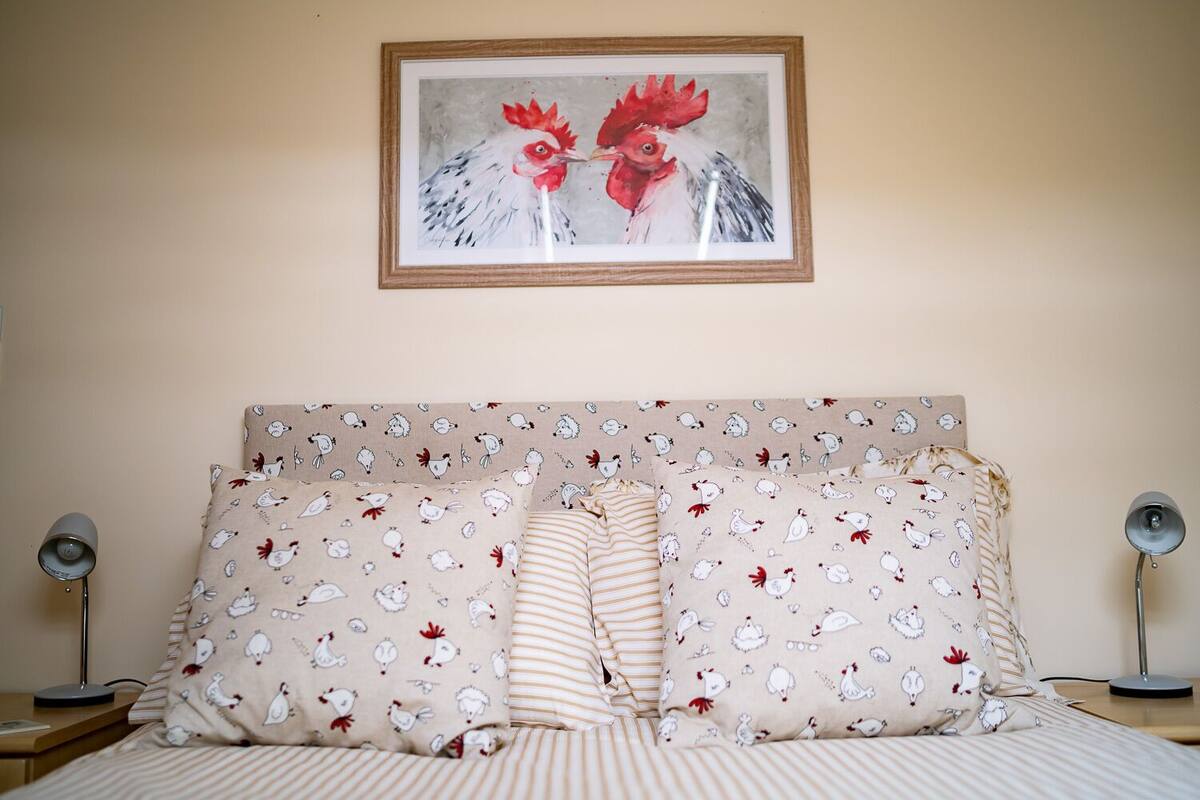 Un dormitorio con una cama, ropa de cama a rayas, un cabecero con una pintura de gallo y almohadas con estampado de gallina.