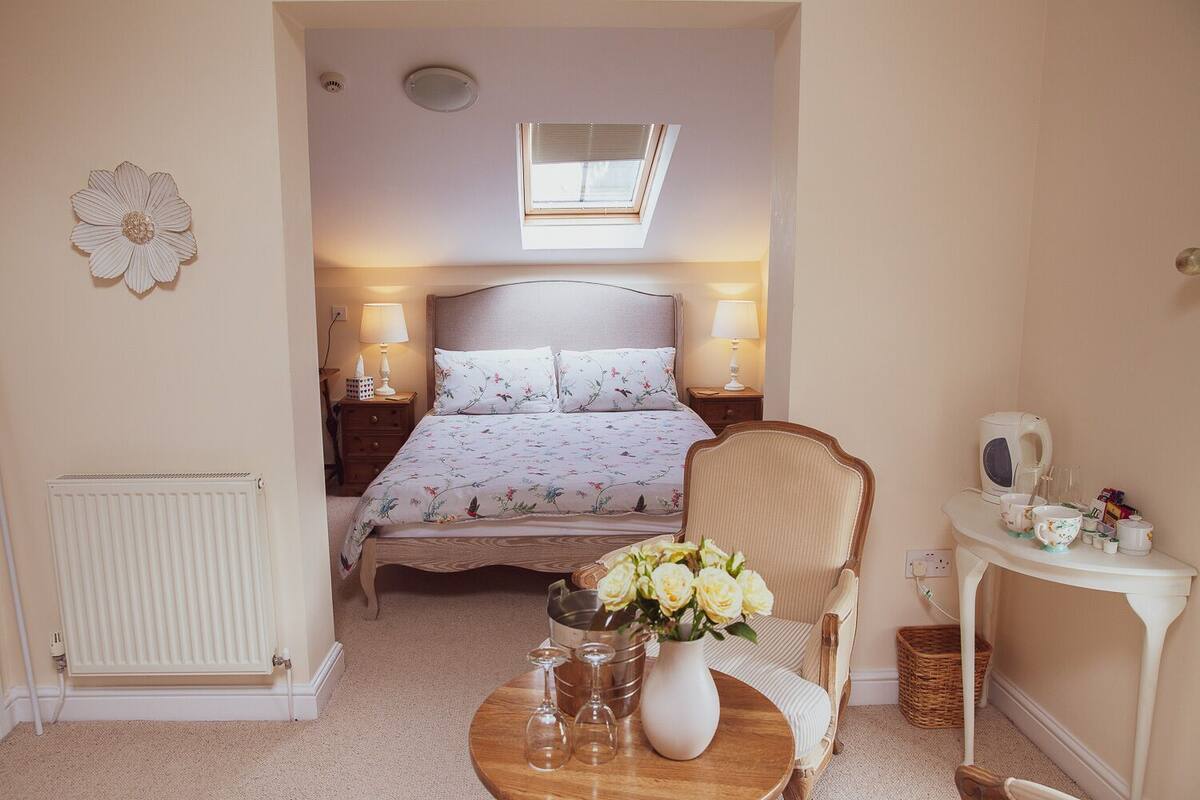 Un dormitorio con cama, mesitas de noche, una silla, una pequeña mesa con un florero y vasos, un radiador, una ventana y una lámpara en el techo.