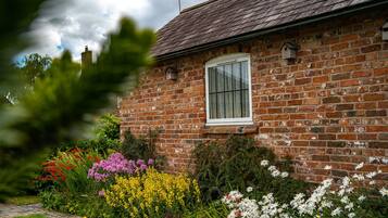 Una casa de ladrillo con ventana, rodeada por un jardín con diversas flores y un sendero.