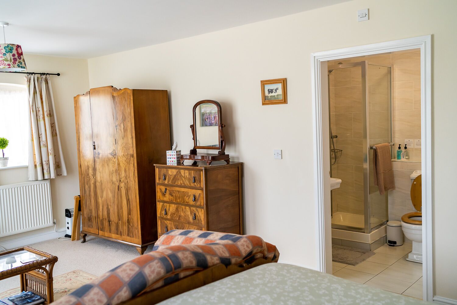 Un dormitorio con cama, armario de madera, cómoda, espejo, ventana con cortinas y baño con ducha e inodoro.