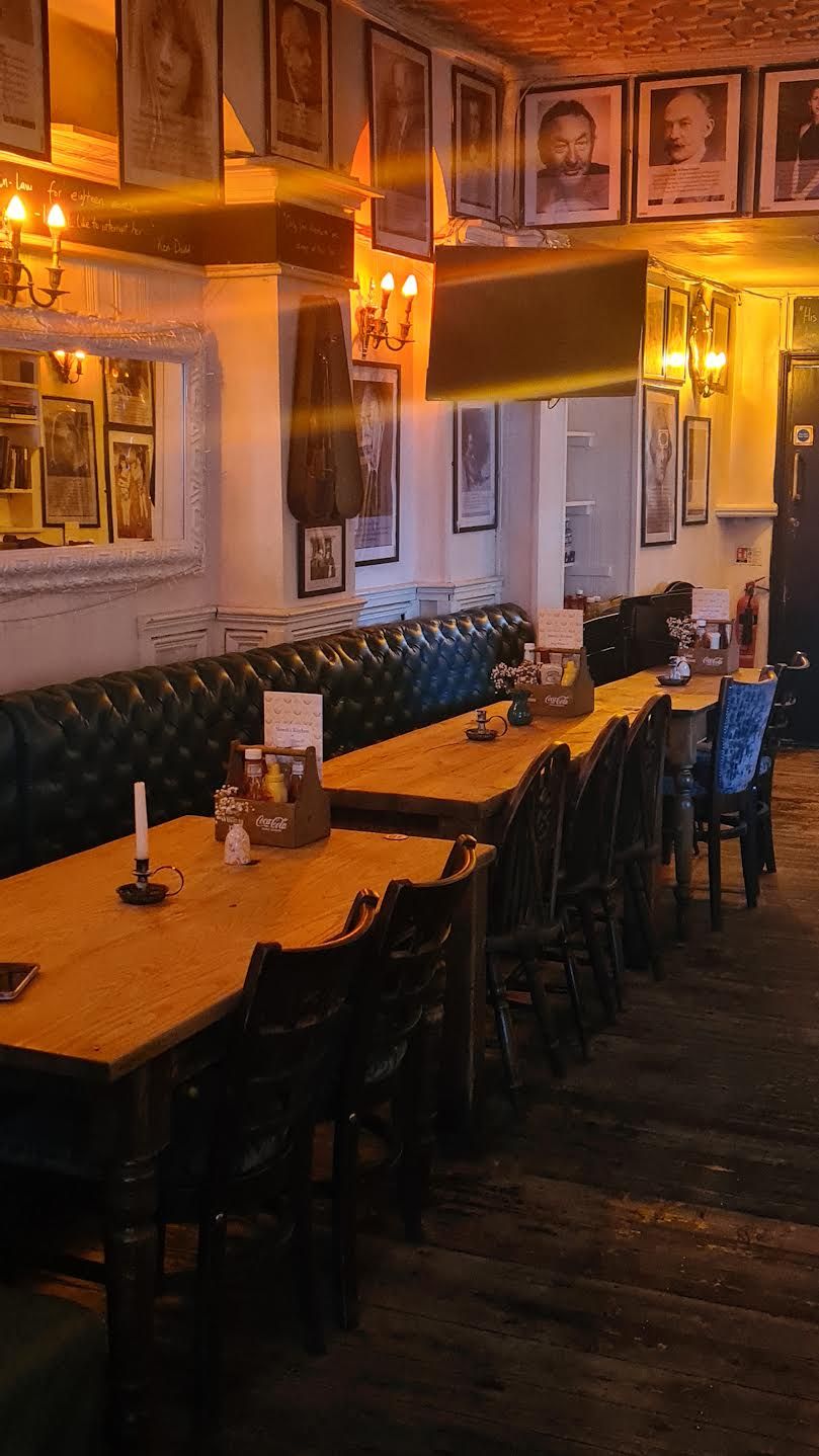 Un interior de restaurante con mesas de madera, asientos tipo banqueta de cuero negro y cuadros en las paredes.