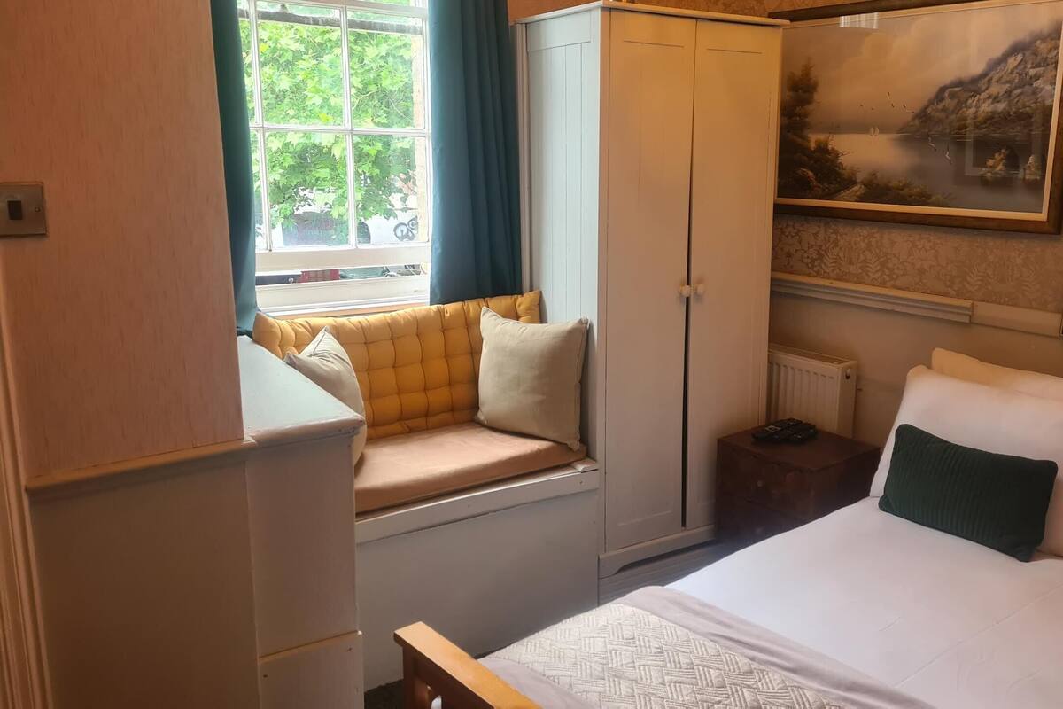 A bedroom with a bed, a window seat with cushions, a wardrobe, and a painting on the wall.
