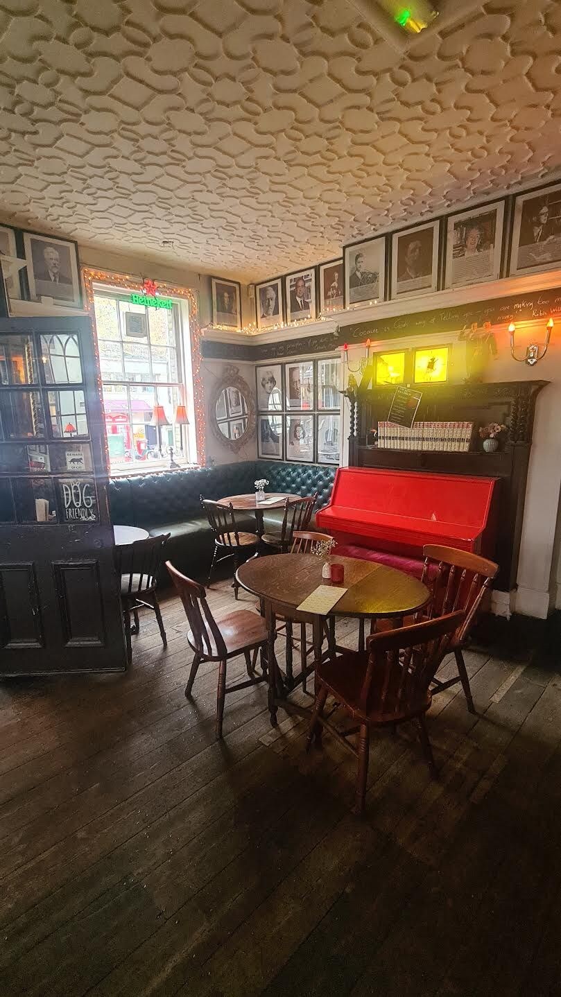 A restaurant interior with a wooden floor, round tables, and chairs. There is a red piano and a bar area with a counter.