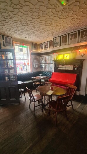 L’intérieur d’un restaurant présente un sol en bois, des tables rondes et des chaises. On y trouve un piano rouge et un coin bar avec un comptoir.