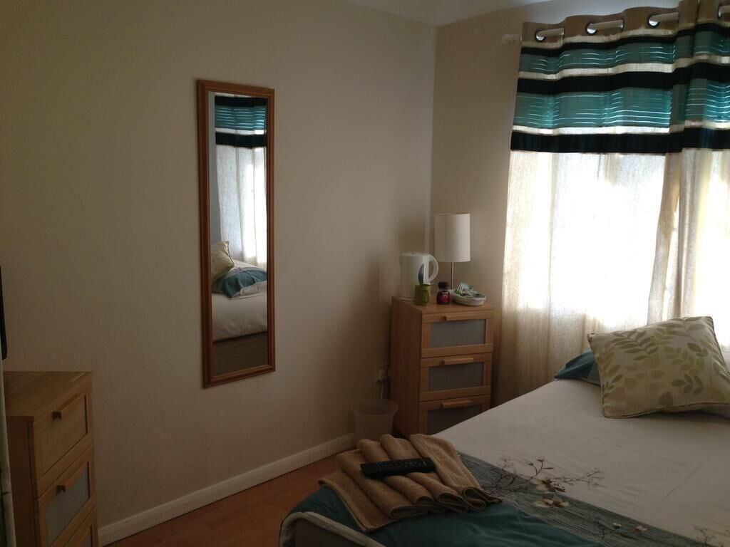 A bedroom with a bed, a bedside table with a lamp, a mirror, and a window with curtains.