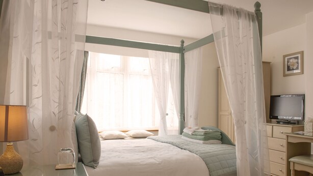 Un dormitorio con cama con dosel, una mesita de noche con lámpara, un televisor sobre un tocador y una ventana con cortinas traslúcidas.