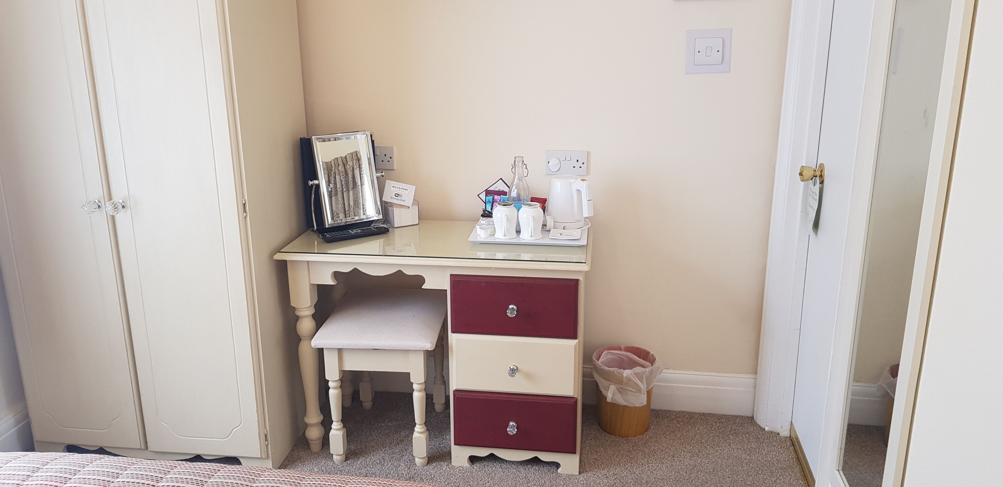 Une petite chambre à coucher, bien rangée, avec un bureau blanc, une chaise, un miroir et une petite poubelle.