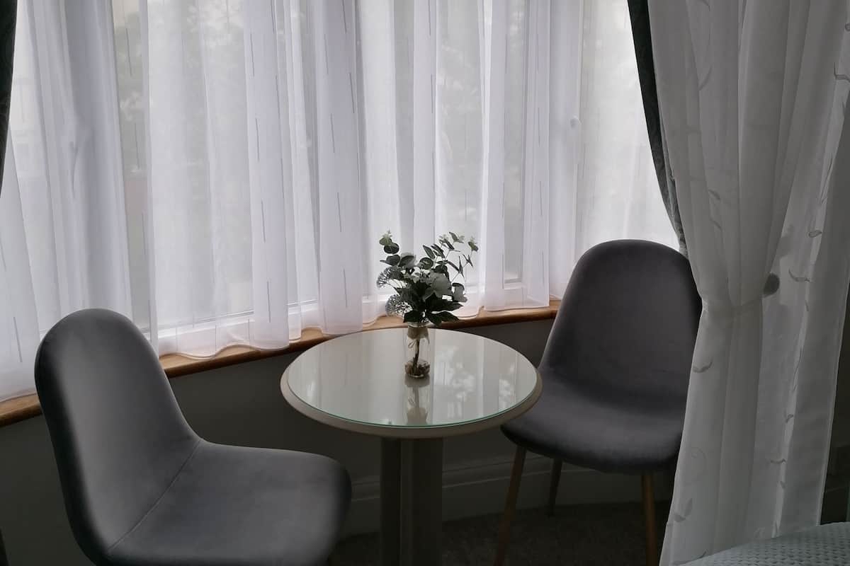 A room with a bed, a round glass table, two chairs, and a window with sheer curtains.