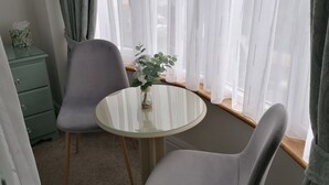 Una habitación con una mesa redonda de vidrio, dos sillas grises, un pequeño jarrón con una planta y una mesita de noche verde con un dispensador de pañuelos.