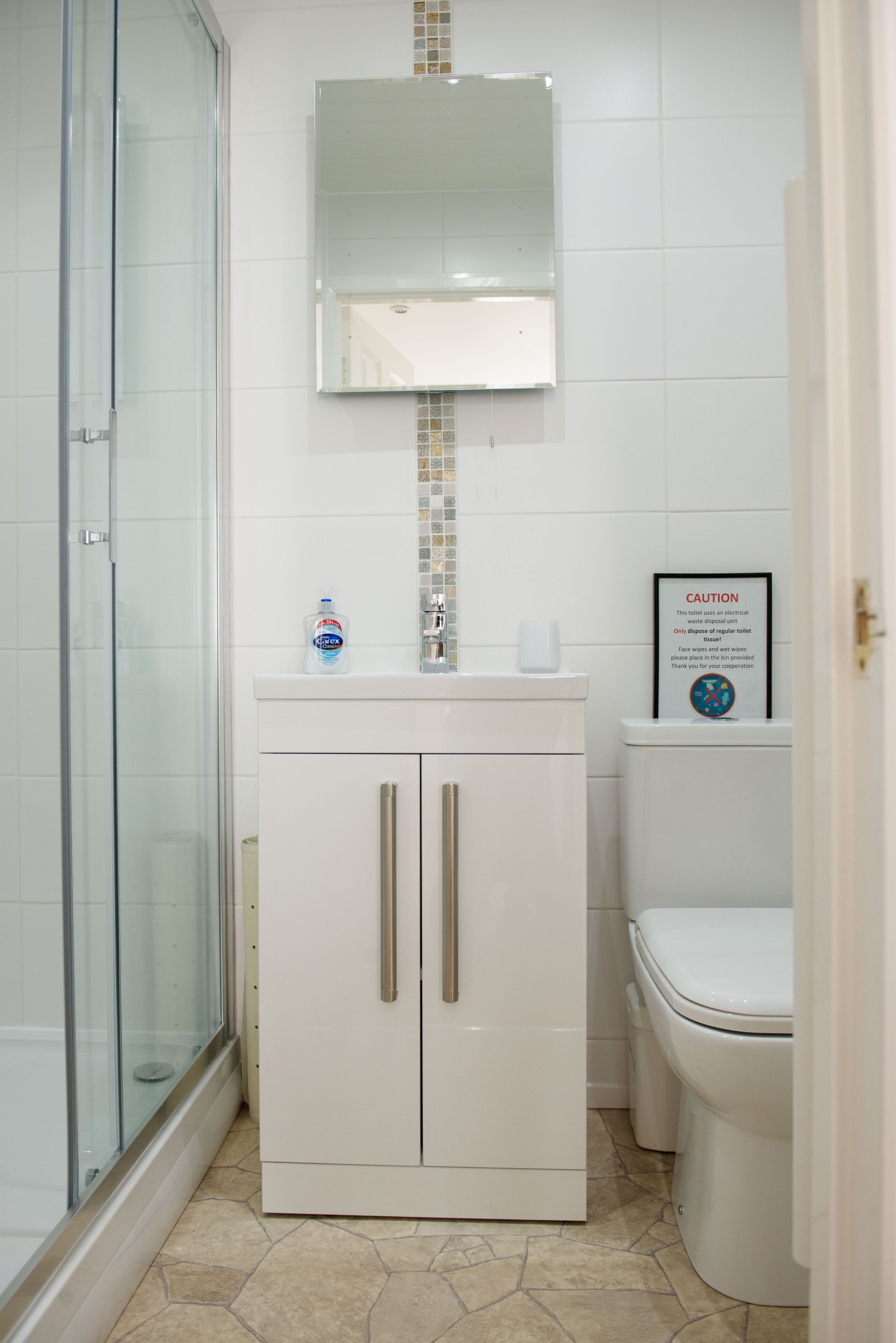 A bathroom with a glass shower enclosure, a white sink with a mirror above, and a toilet with a caution sign.