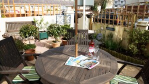 Una mesa redonda de madera con una revista, una botella y un vaso. Hay dos sillas con cojines a rayas. El entorno incluye plantas en macetas y una valla de madera.