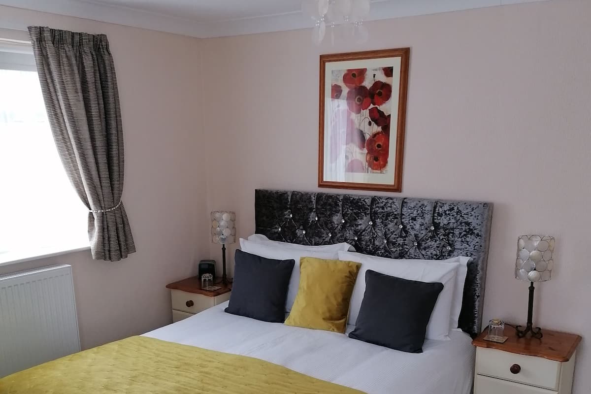 A bedroom with a bed, a bedside table, a nightstand, a lamp, a framed picture, and a window with curtains.